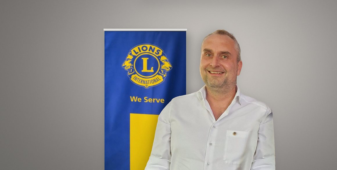 Alexander Maintok, Präsident des Lions Club Augsburg-Friedensstadt 2026/2027, im weißen Hemd vor dem blau-gelben Lions-Banner mit dem Emblem von Lions International und dem Leitspruch „We Serve“.