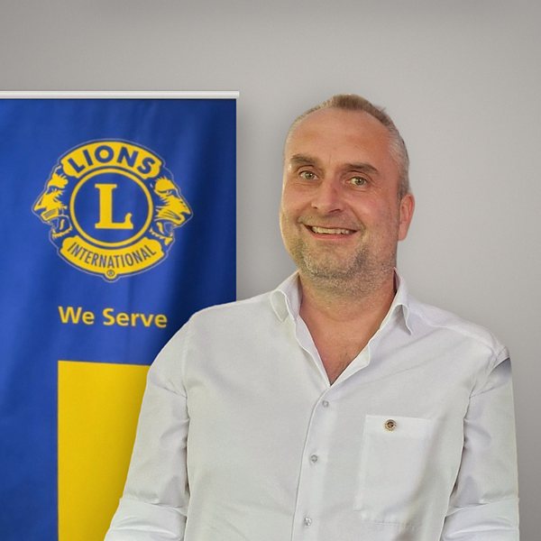 Porträt von Alexander Maintok im weißen Hemd, Präsident des Lions Club Augsburg-Friedensstadt im Clubjahr 2026/2027. Hinter ihm das Lions-Banner mit dem Emblem von Lions International und dem Leitspruch „We Serve“.