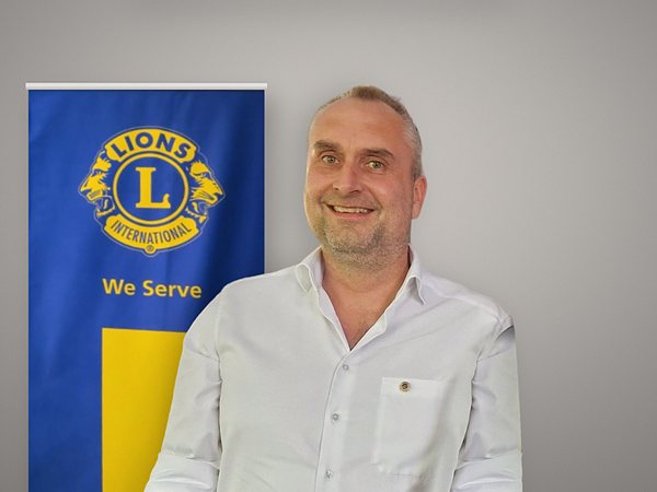 Pressefoto im Querformat: Alexander Maintok im weißen Hemd vor dem Lions-Banner mit Emblem.